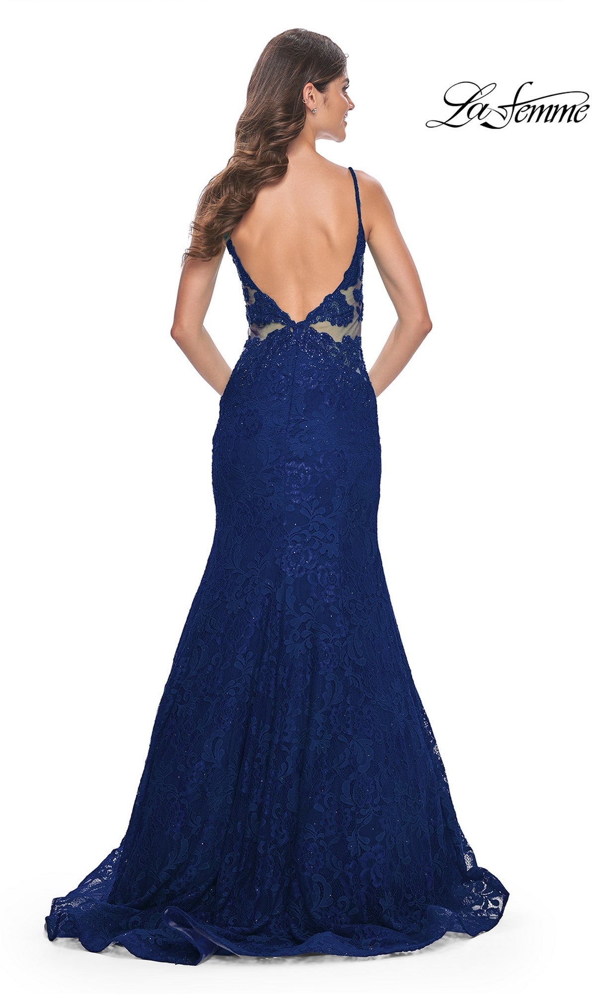 A woman wears the La Femme Sheer-Sides Long Lace Prom Dress 32315, a floor-length dark blue mermaid gown with thin straps and a low open back, standing and facing away from the camera.